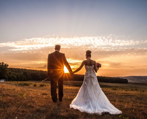 Ein Hochzeitsfotograf fängt ein Brautpaar Händchen haltend auf einem Feld bei Sonnenuntergang ein.