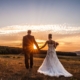 Ein Hochzeitsfotograf fängt ein Brautpaar Händchen haltend auf einem Feld bei Sonnenuntergang ein.