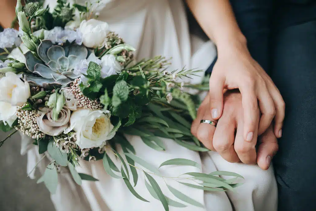 Die Hände einer Braut und eines Bräutigams, die einen Blumenstrauß halten.