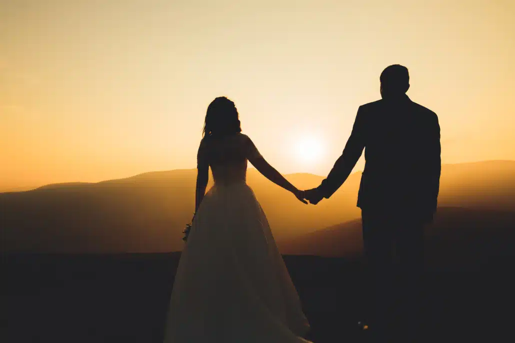 Eine Braut und ein Bräutigam halten sich bei Sonnenuntergang auf einem Berg an den Händen.